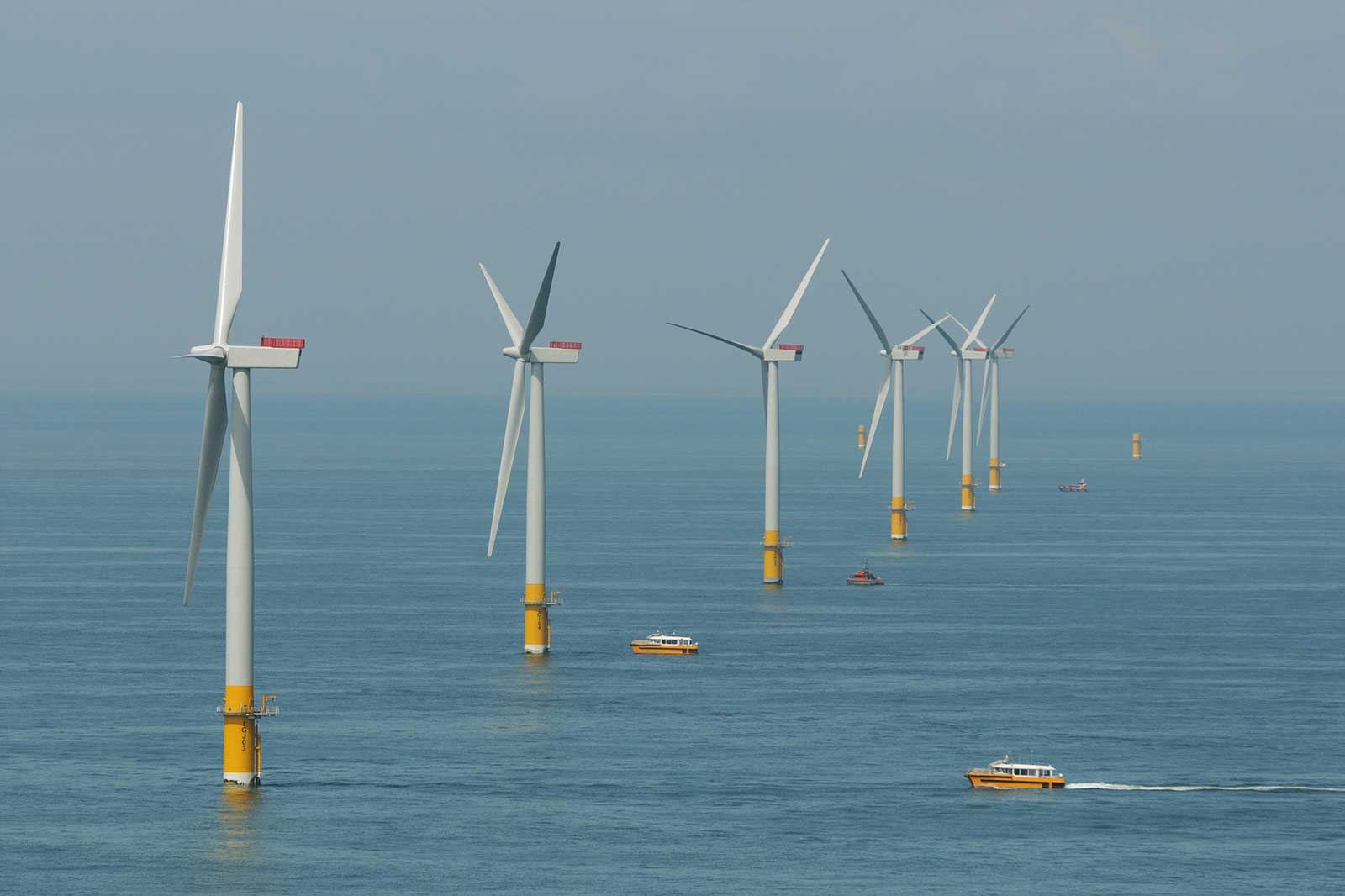 A row of offshore wind turbines in the ocean, with boats navigating nearby. The water is calm and blue under a clear sky.