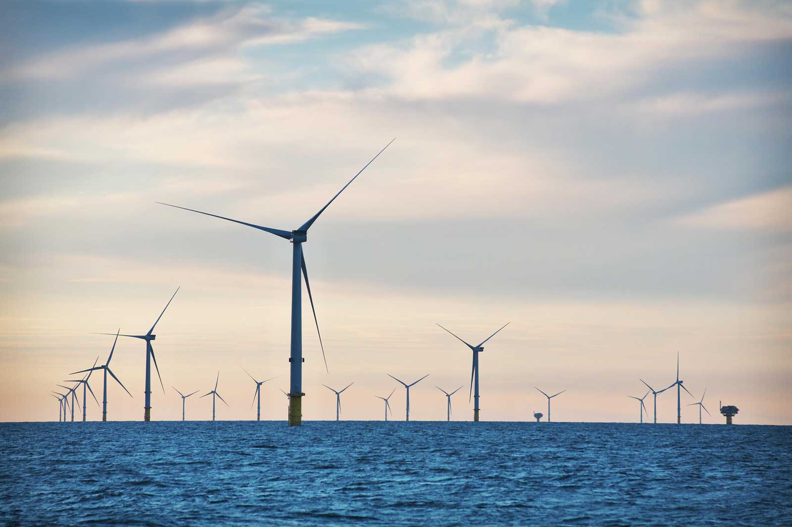 A serene offshore wind farm with multiple turbines on the horizon, set against a soft, cloudy sky and a calm sea.