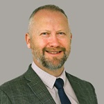 A man in a grey suit with a checkered shirt and black tie poses in front of a grey background.