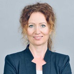A woman wearing a dark jacket poses against a light grey background. She has curly hair and a neutral expression.