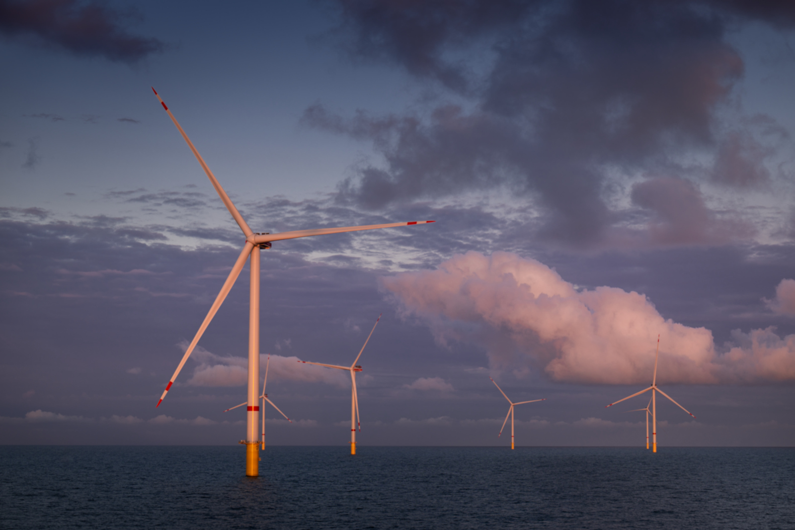 Offshore wind turbines stand in the sea under a colourful sky with clouds at dusk.