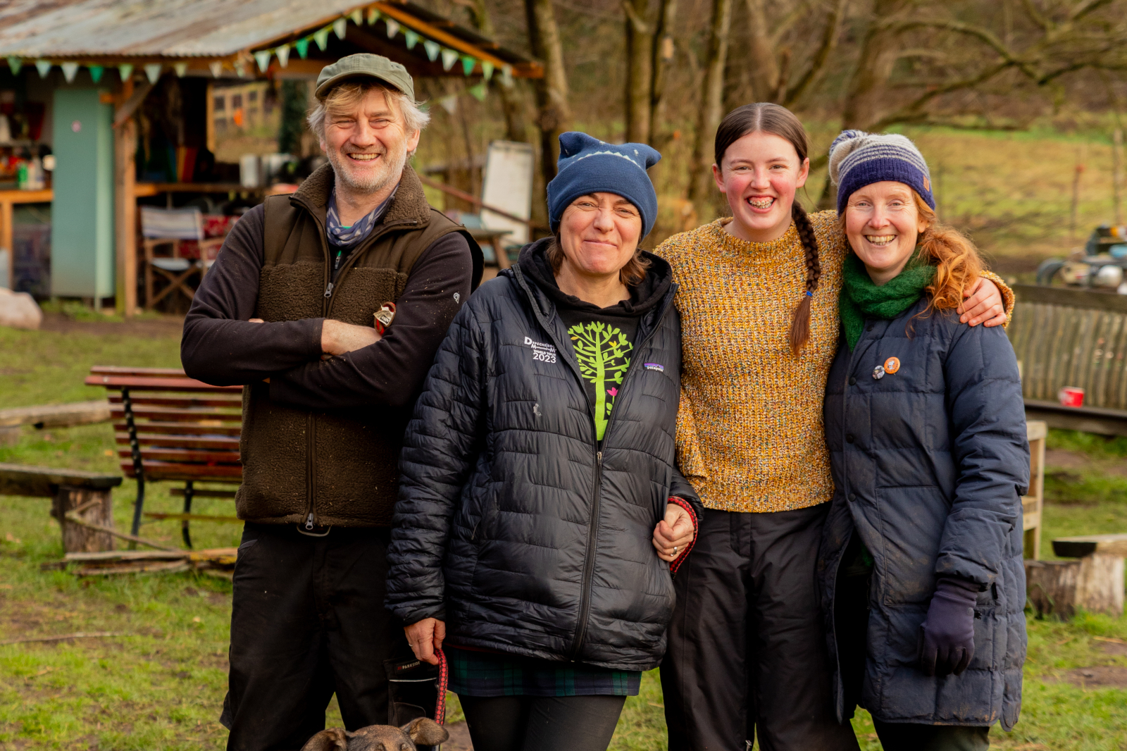 Four people dressed in warm clothing stand outdoors in a rustic setting with wooden benches and a small building in the background.