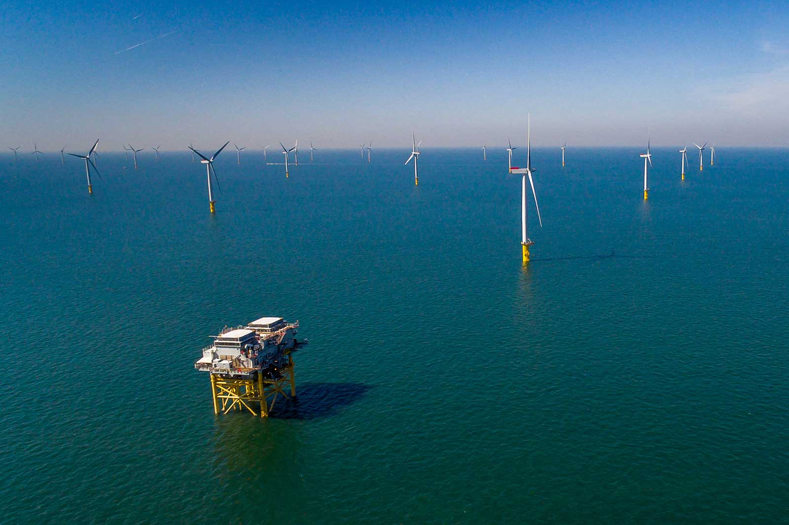An aerial view of an offshore wind farm with a central platform and multiple wind turbines in the blue sea.